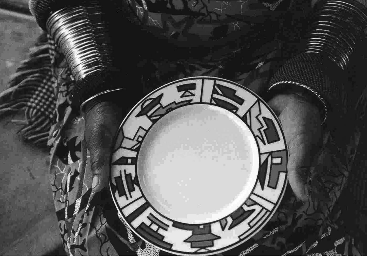 two hands with stacked bracelets, holding a plate towards the camera. The plate has geometric patterns along the rim. 